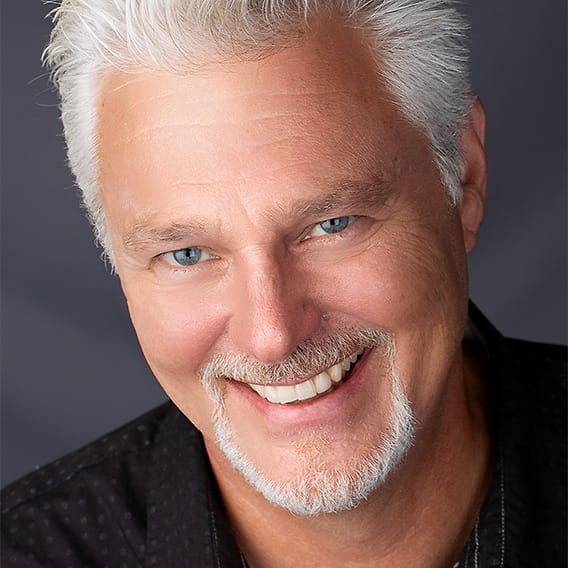 Character headshot of a man with silver hair and a goatee in a black dotted shirt