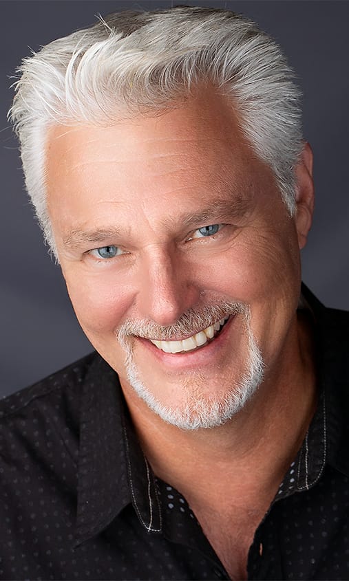 Character headshot of a man with silver hair and a goatee in a black dotted shirt